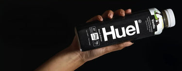 A hand holding a bottle of Huel Vanilla Black Edition against a black background
