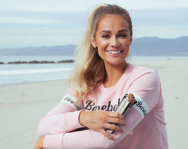 A woman holding a Barebells protein bar on the beach. 