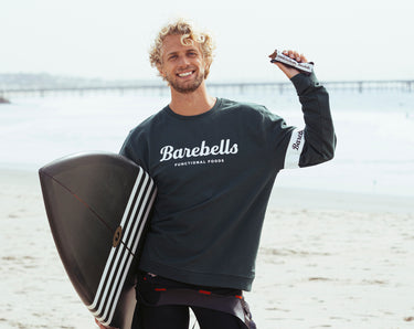 A man holding a Barebells protein bar and a surfboard on the beach.